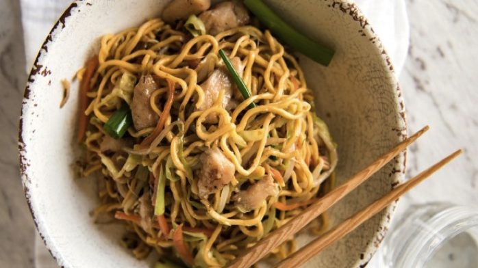 A bowl of brown egg noodles with chicken and vegetables and chopsticks.