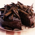 A round chocolate Cake with chocolate peels on top with one piece taken out.