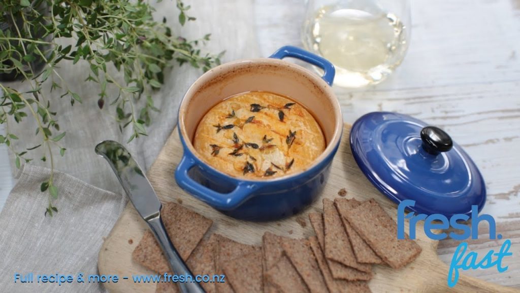 Small blue pot with a whole round cheese in it topped with garlic slices and herb, thyme and glass of wine, crackers around the pot.