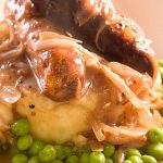 Two sausages and cooked onions covered with gravy on bed of peas and mash.