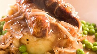 Two sausages and cooked onions covered with gravy on bed of peas and mash.
