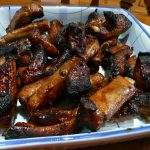 Dark brown pork spare ribs heaped in a blue rimmed square bowl.