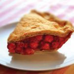 A piece of cherry pie on a plate on pink striped table cloth.