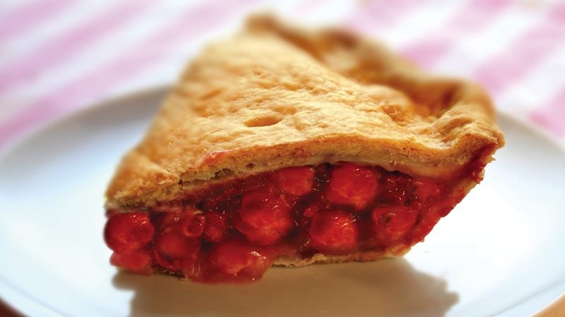 A piece of cherry pie on a plate on pink striped table cloth.