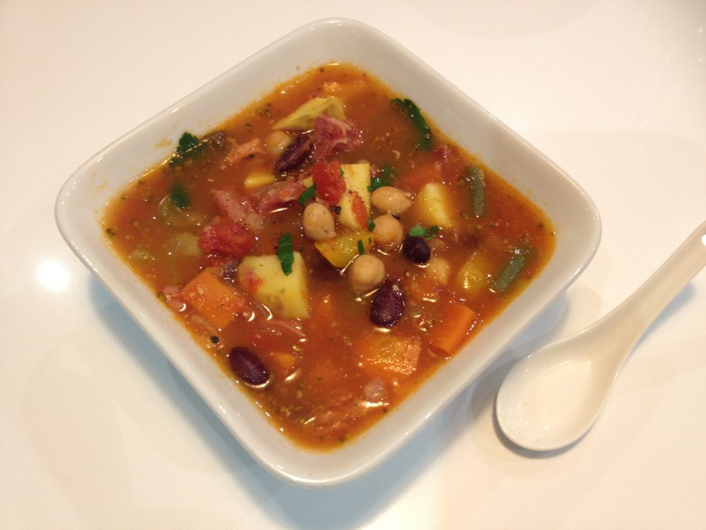 A square bowl of red soup with lots of ingredients and a spoon.