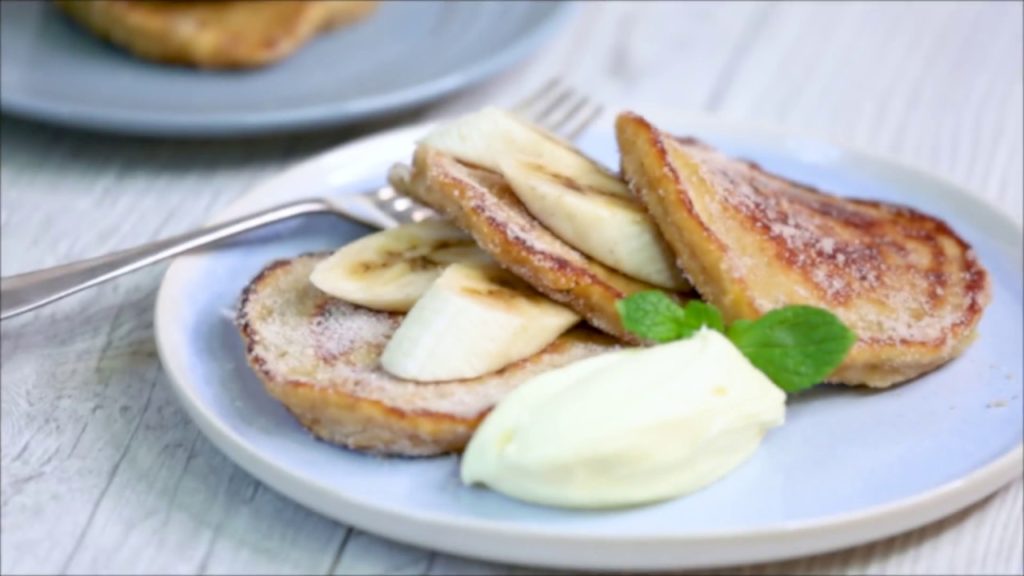 Three round pancakes with banana slices inserted in between and a blob of white cream on blue plate with a fork.