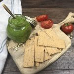 A jar of green pesto, crackers and tomatoes on wooden board.