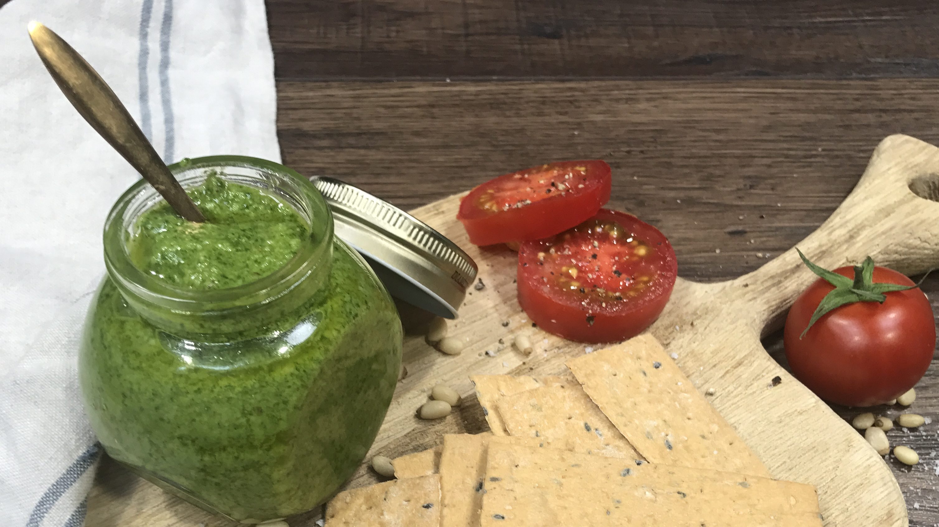 A jar of green pesto, crackers and tomatoes on wooden board.