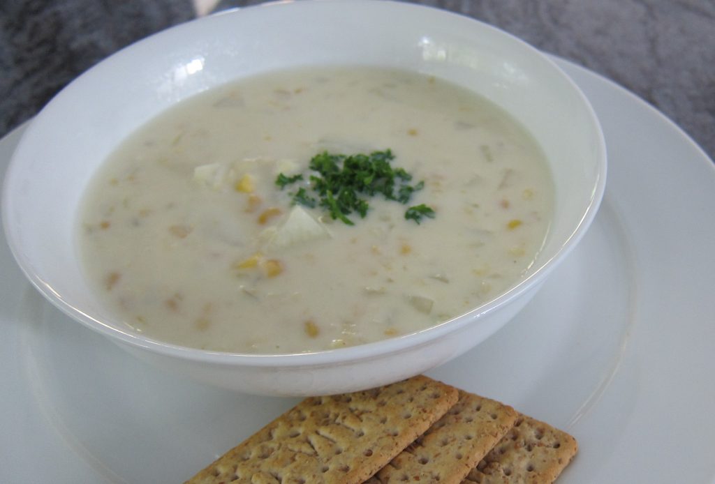 A bowl of whitish soup with corn and parsley on top, a few crackers at side.