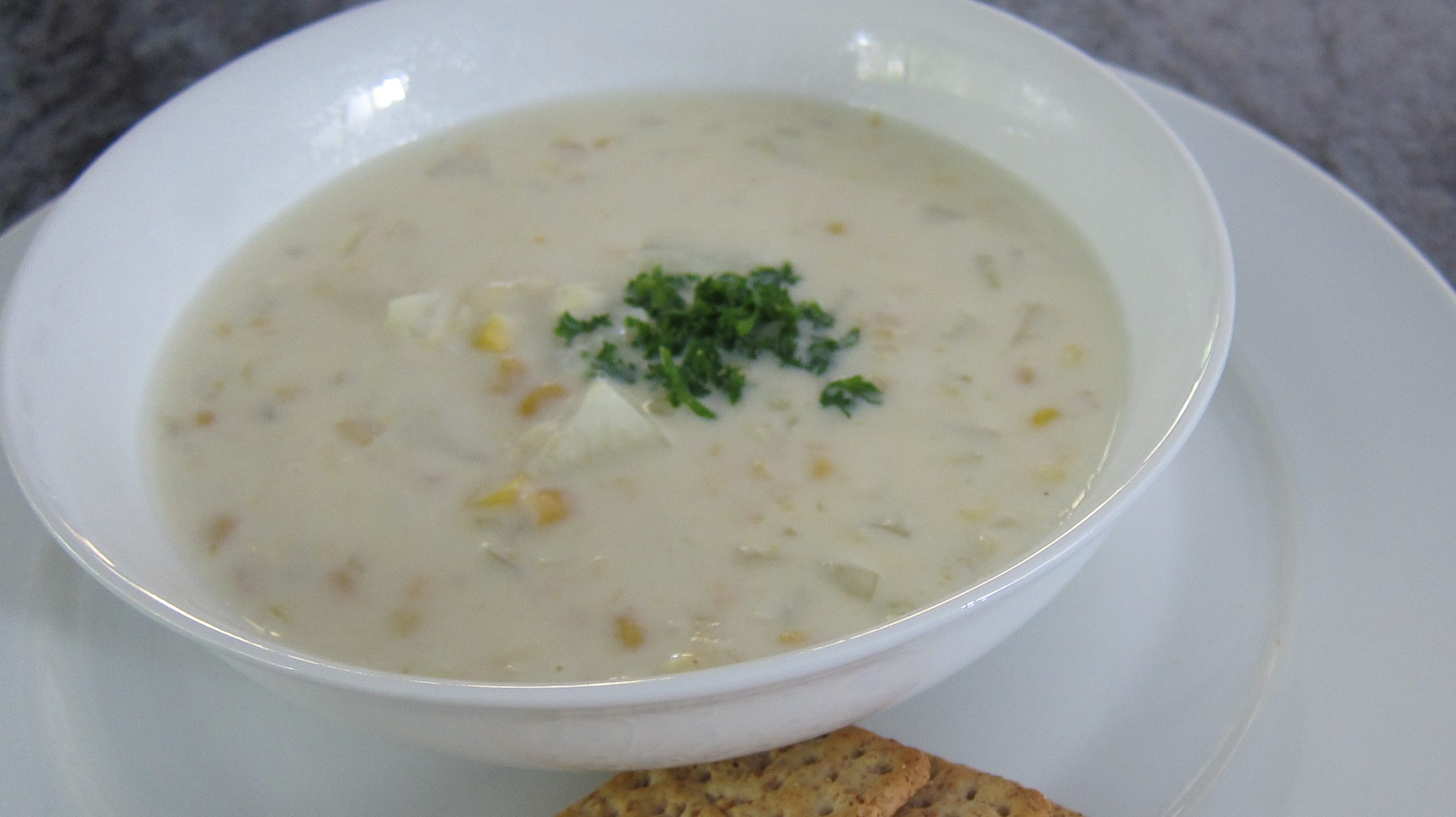 A bowl of whitish soup with corn and parsley on top, a few crackers at side.