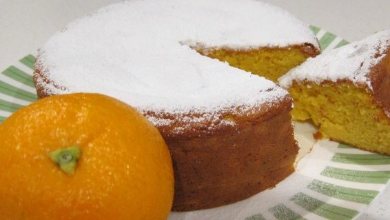 A round whole cake with one slice being served and an orange in the front.