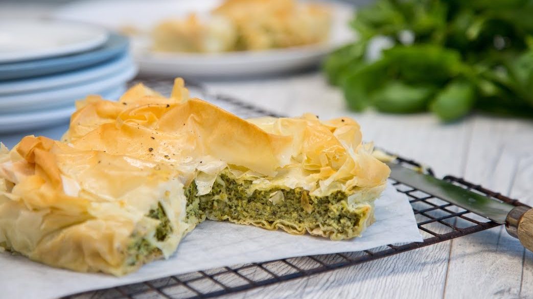 A whole green filling filled pie, a slice cut out on wire rack and plates stack in the back.