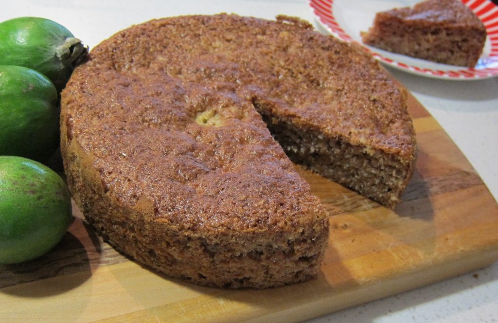 A whole brown cake, slice taken out and three green fruit on left.