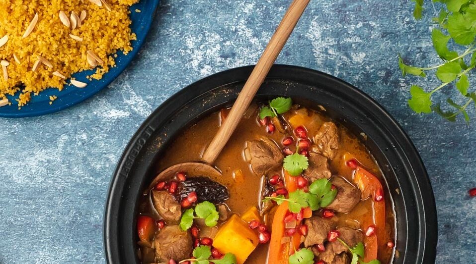 Black pot full of red stew with meat and potato topped with red berries and herbs and a plate of yellow couscous on blue top