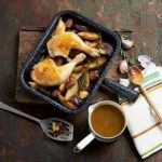 Two cooked chicken legs, several potatoes and sausages in a black roasting tin, serving utensils and brown gravy in a jug.