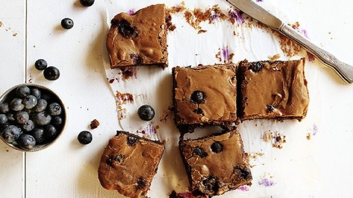 Five square pieces of brownies and a pot of blueberries on white board.