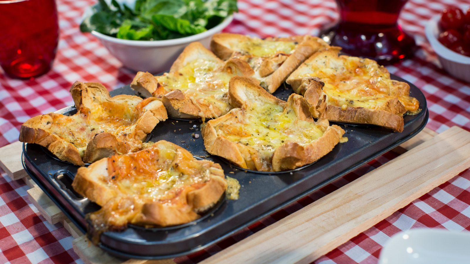 Six hole muffin tin full of bread cups, green salad on red & white tablecloth with red glasses.
