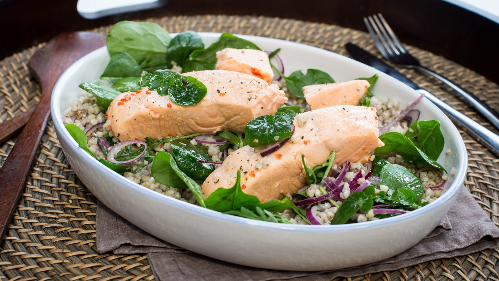Two cooked pieces of salmon on grain and green salad in oval bowl, on a tray with forks.