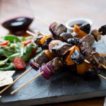 Six meat and vegetable skewers stack on stone slate with pieces of flat bread.