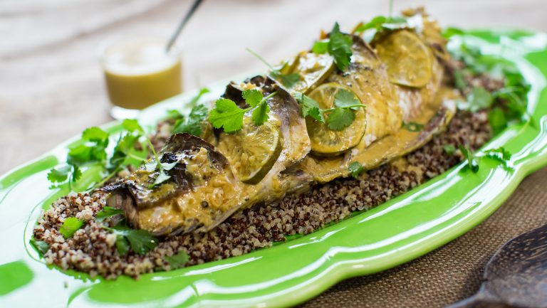 A whole cooked fish topped with yellowish sauce, lemon slices and green herbs on bed of wholegrain mustard on a light green platter, a jar of sauce in the back.
