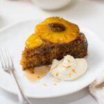 A piece of brown cake with a pineapple ring on top on a white plate with a blob of cream and fork.