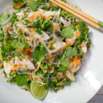 Rice noodle and green salad with red chilli bits and lime halves and chopsticks on white round plate