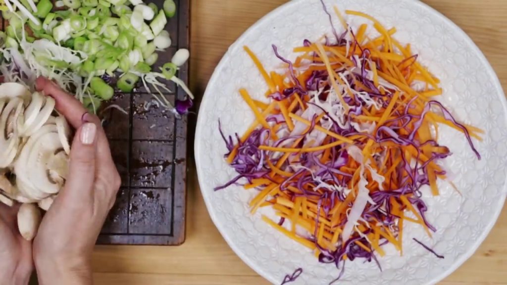 A white plate with carrot and red cabbage slices heaped, two hands holding full of mushroom slices on left of the plate.