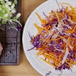 A white plate with carrot and red cabbage slices heaped, two hands holding full of mushroom slices on left of the plate.