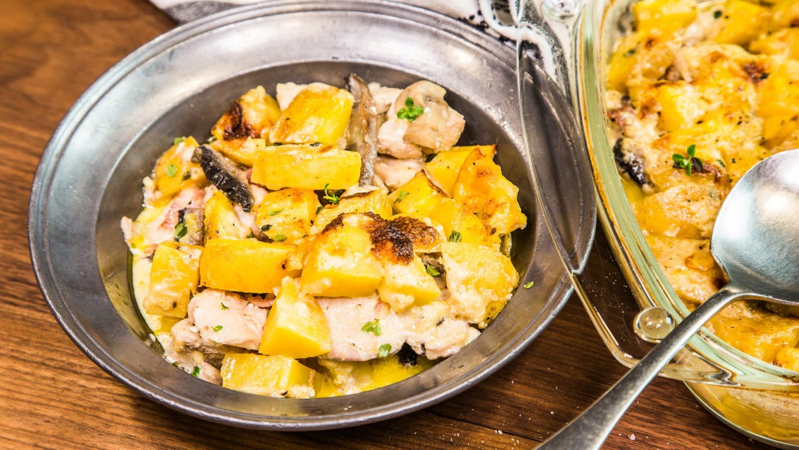 A serving bowl and a glass oven baking dish full of yellow potato bake with a spoon.
