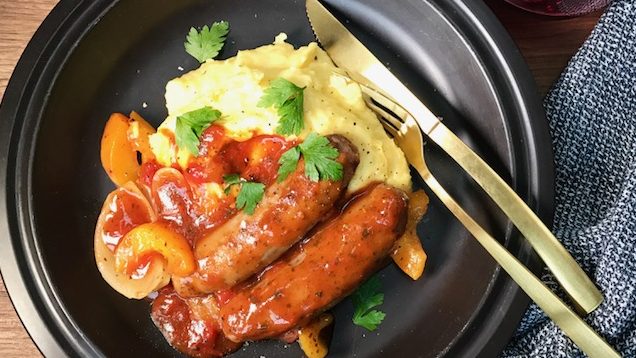Two sausages in red tomato sauce on mashed potato and peas on a black plate with a knife and fork.