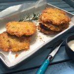 Five round hash browns on paper in a metal tray with sprig of rosemary, and a knife.