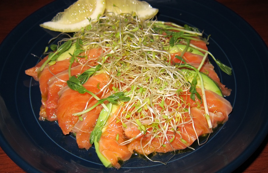 salmon slices and avocado slices toppd with sprouts and a lemon wedge on a plate.