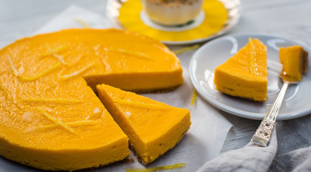Orange coloured round cake, sliced in. A slice on a small plate with a fork. A tea cup saucer in the back.
