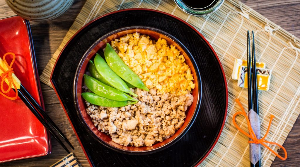 A black round tray with a bowl of eggs, mince and green beans on bamboo mat. chopsticks, small bowl of sauce.