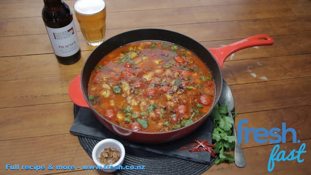 A red skillet full of red curry on brown table with beer.