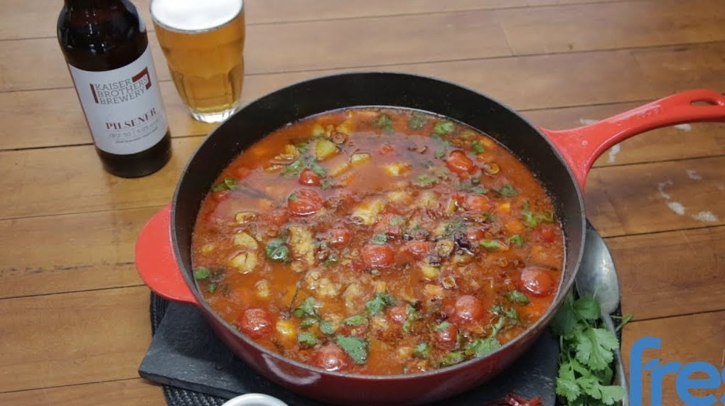 A red skillet full of red curry on brown table with beer.