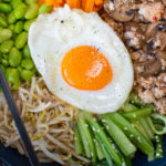 Bowl of chicken, vegetables and a fried egg topped food with chopsticks
