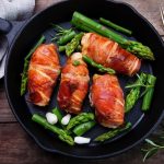 Four bacon wrapped chicken pieces and asparagus on skillet with a fork and pale blue cloth on wooden table.
