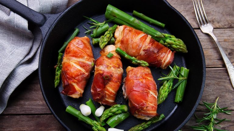 Four bacon wrapped chicken pieces and asparagus on skillet with a fork and pale blue cloth on wooden table.