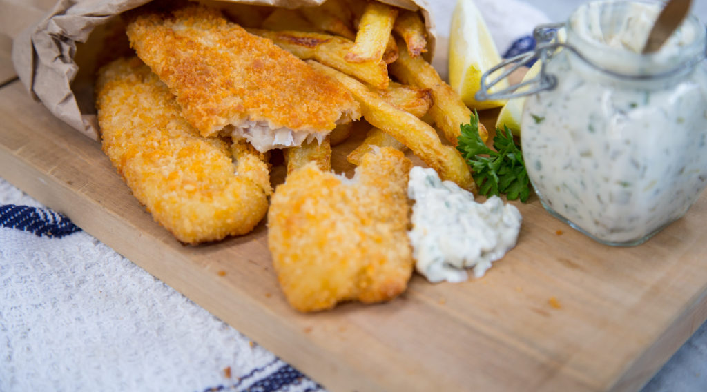 Crumbed fish pieces and chips and jar of tarter sauce on wooden board