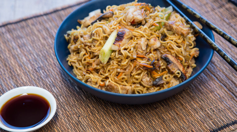 A bowl full of fried noodles on brown bamboo mat with small dish of soy sauce.