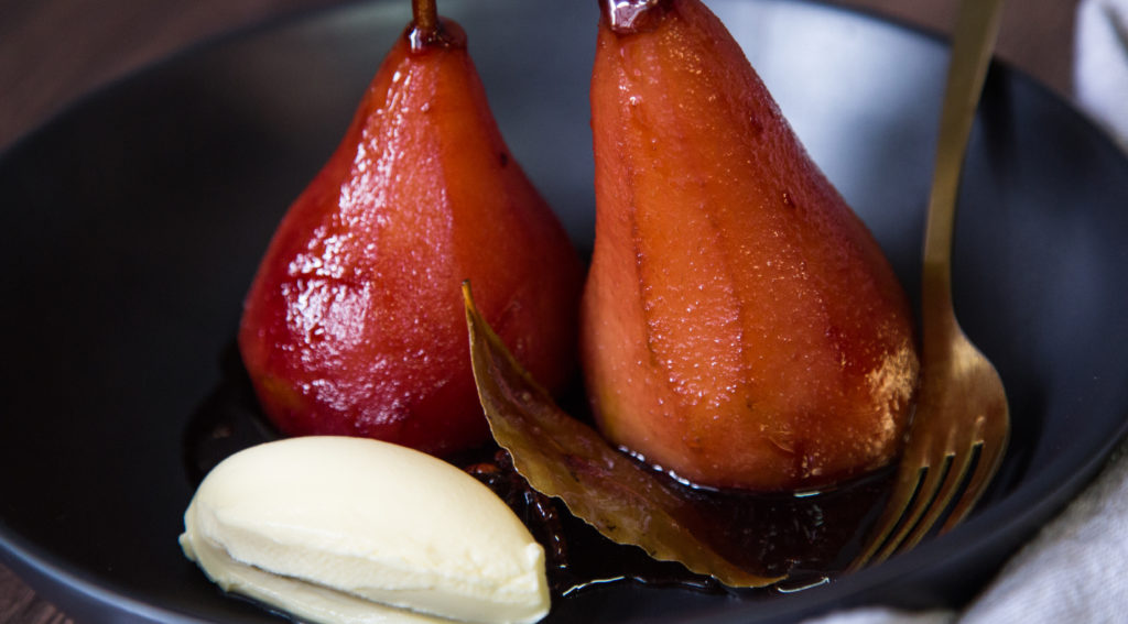 Two whole red poached pears, bay leaf and white cream on a plate.