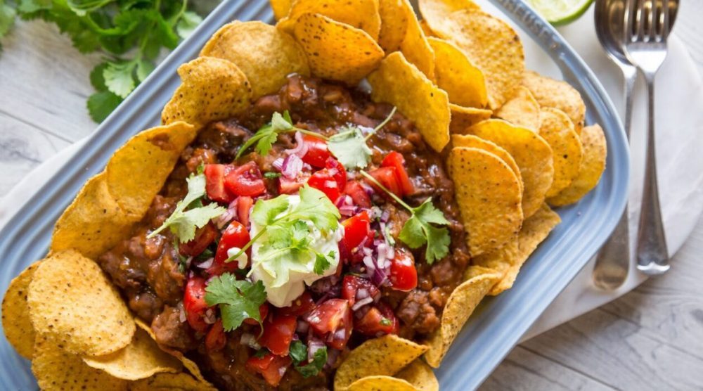 Round corn chips on a platter topped with beef, sauce, red cupsicum pieces, green herb and green mash. fork and spoon on side