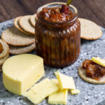 A jar of brown chutney, a block of cheese and some round crackers on a round stone board.