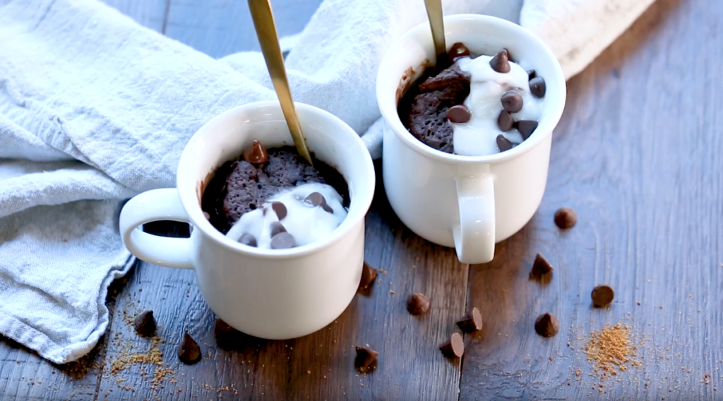 2 white mugs of chocolate cake topped with white cream and chocolate chips and spoons on a table, scattered with chocolate chips and a white cloth