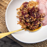 A bowl of fruit crumble with pink yoghurt in white bowl with gold spoon