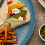 Two wraps filled with brown and red food and white sauce on blue plate with chips, small bowl of capers and a bowl of extra chips.