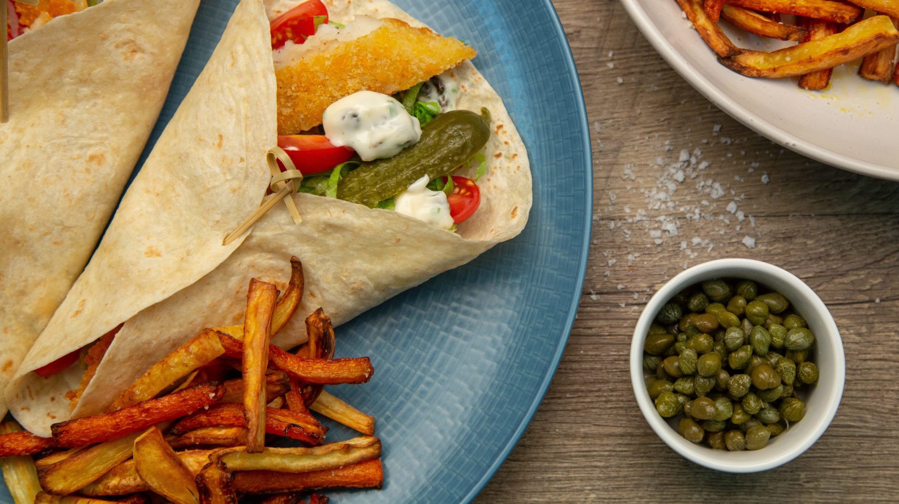 Two wraps filled with brown and red food and white sauce on blue plate with chips, small bowl of capers and a bowl of extra chips.