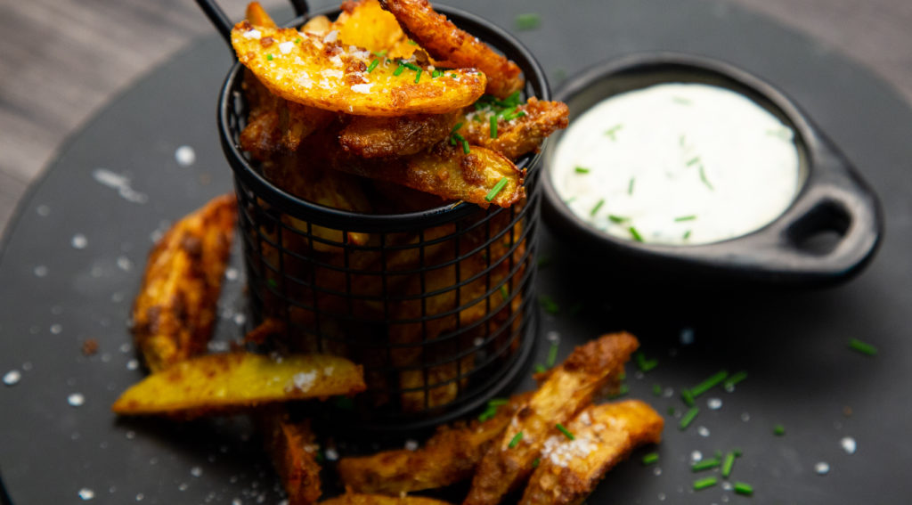 A basket of brown potato wedges and a small dish of white sauce on a black plate.
