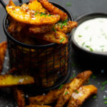 A basket of brown potato wedges and a small dish of white sauce on a black plate.